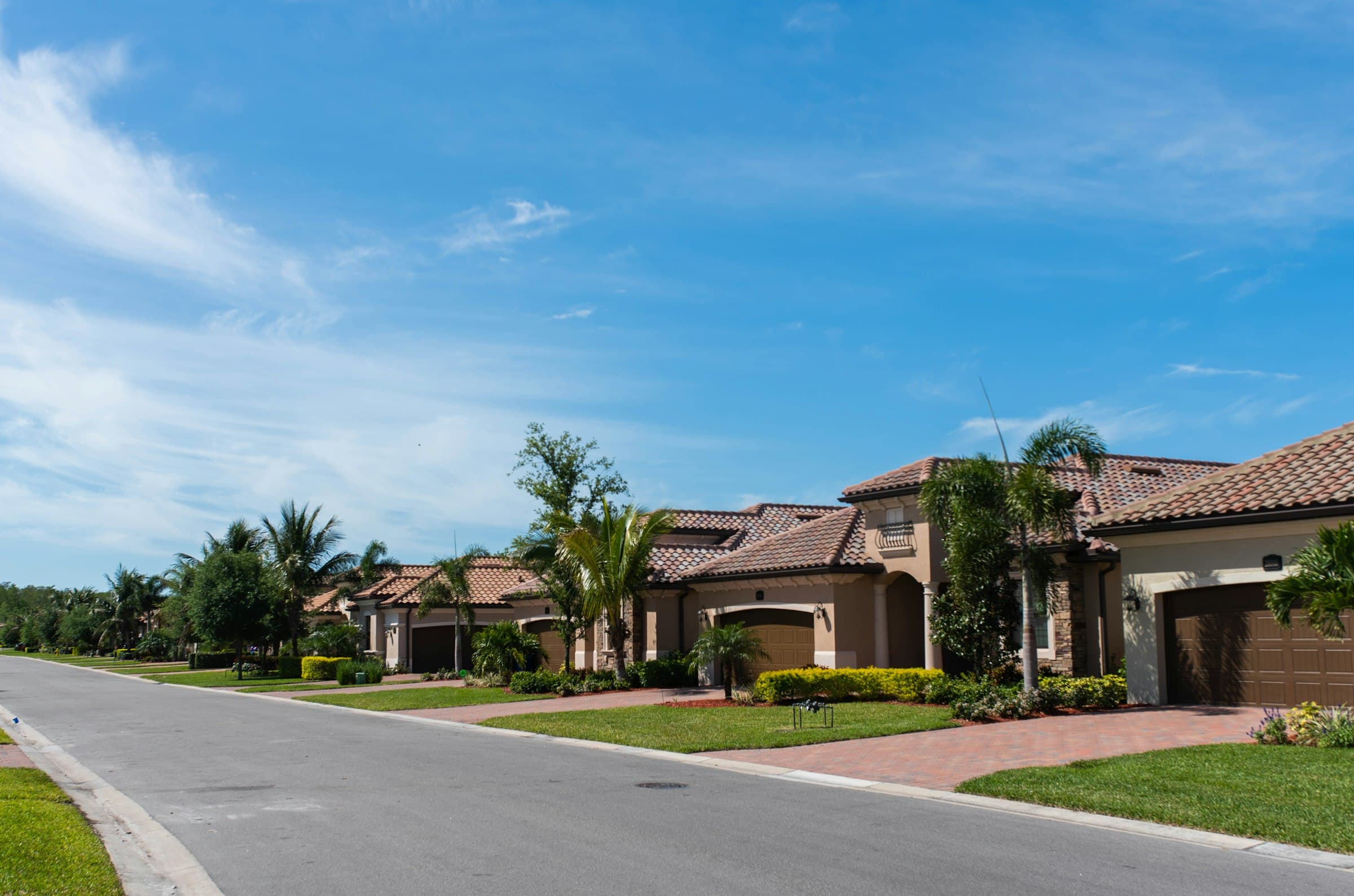 A residential street of stucco homes with barrel-tile roofs, palm trees and clear sky