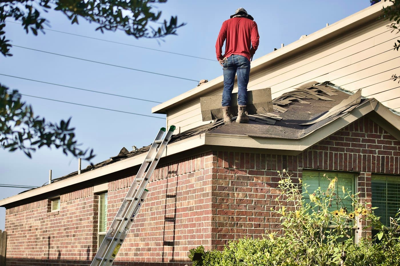 A worker standing on a partly stripped roof during a tear-off, ladder against the wall